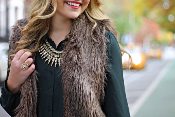 Woman wearing a brown faux fur vest over a dark green blouse with a gold statement necklace