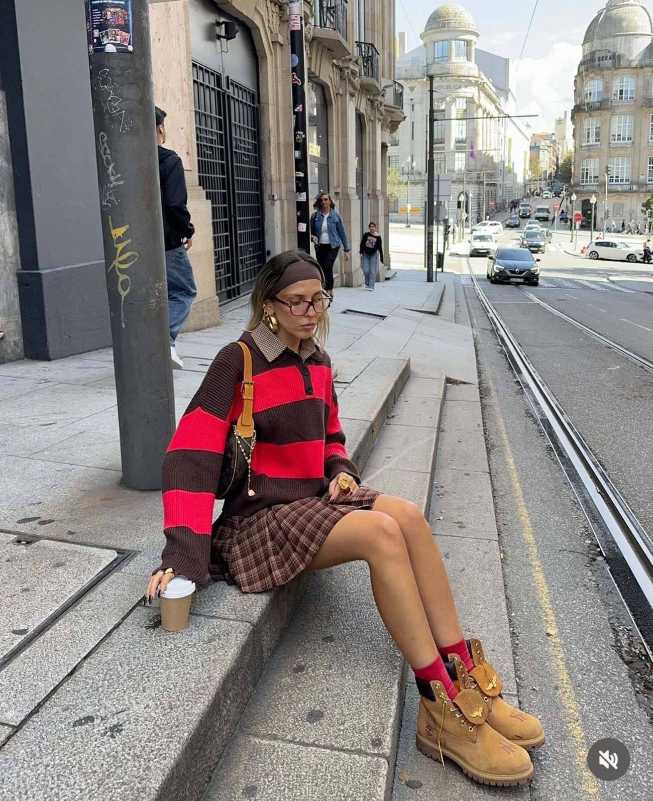 Woman in a red and brown striped knit polo sweater, brown plaid pleated mini skirt, pink socks, and wheat Timberland boots
