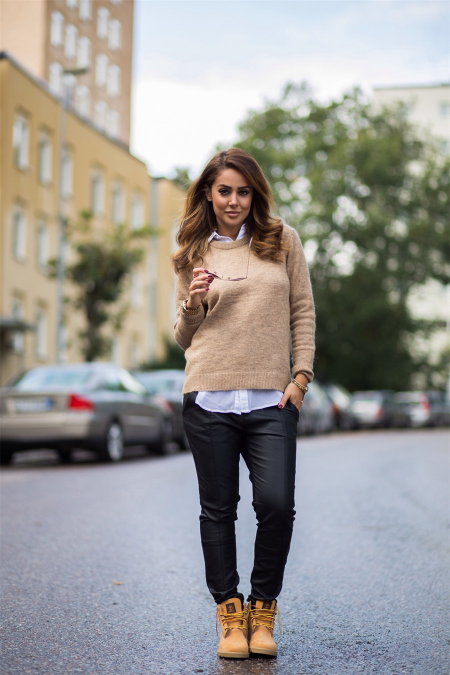 Woman wearing a camel knit sweater over a white collared shirt, black leather-look skinny pants, and wheat Timberland boots