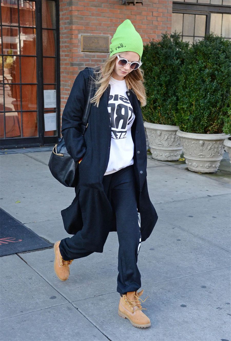 Woman in a neon green beanie, white graphic sweatshirt, black coat, black sweatpants, and wheat Timberland boots