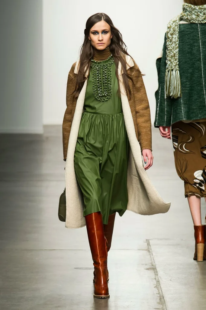 Runway model in a forest green dress, cream shearling coat and rust-brown knee boots