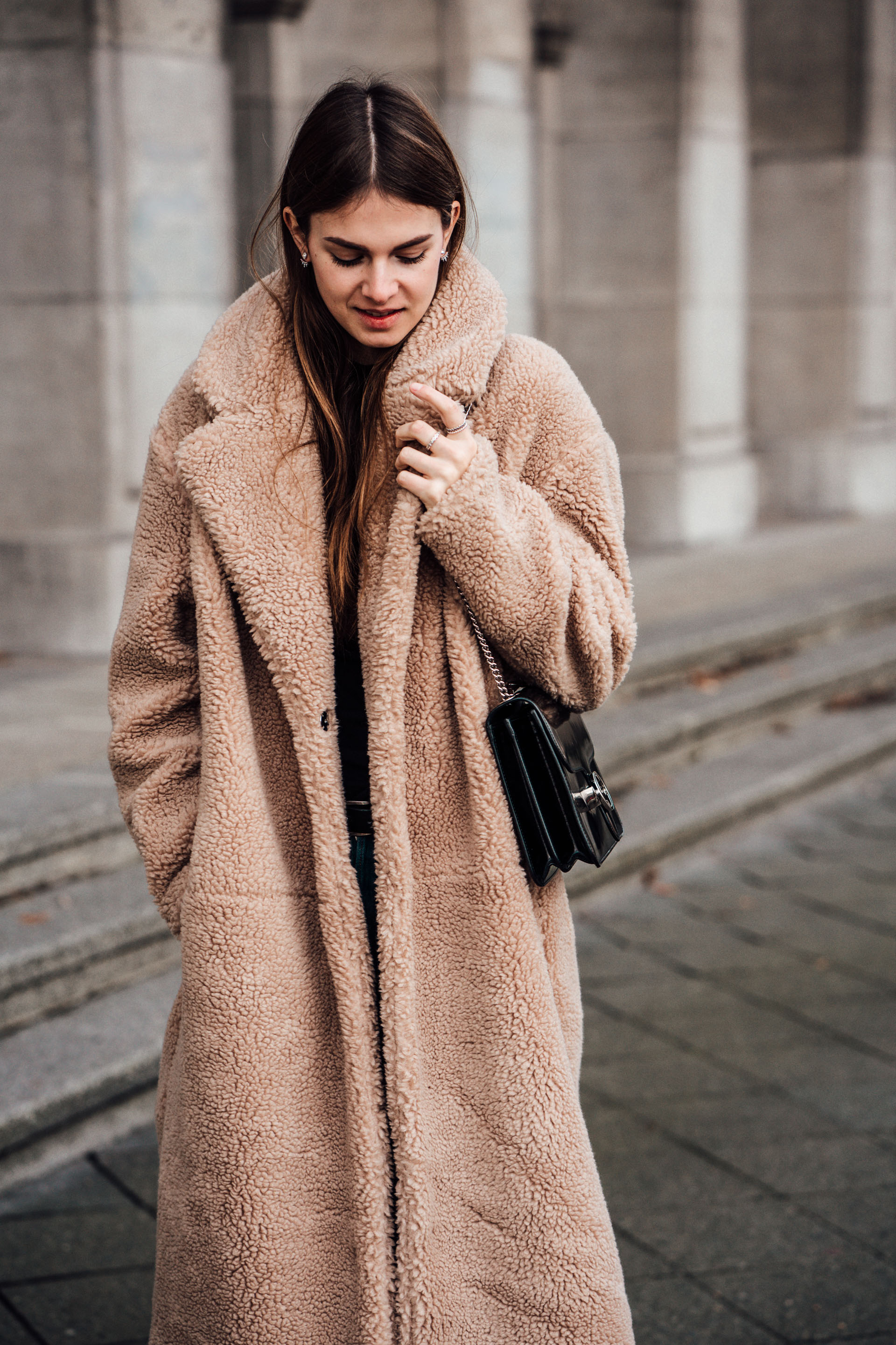 Woman wearing a long beige teddy coat with a small black chain-strap bag