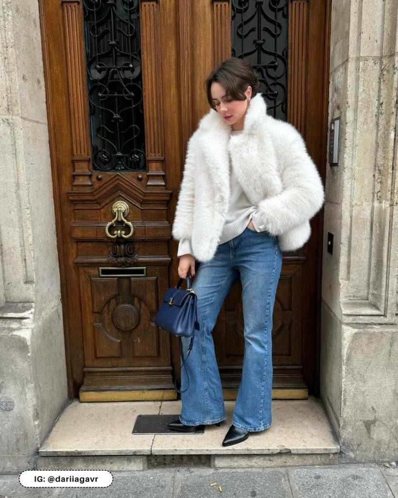 A white cropped faux fur jacket worn over a white top with blue flared jeans, a navy mini bag and black pointed boots.