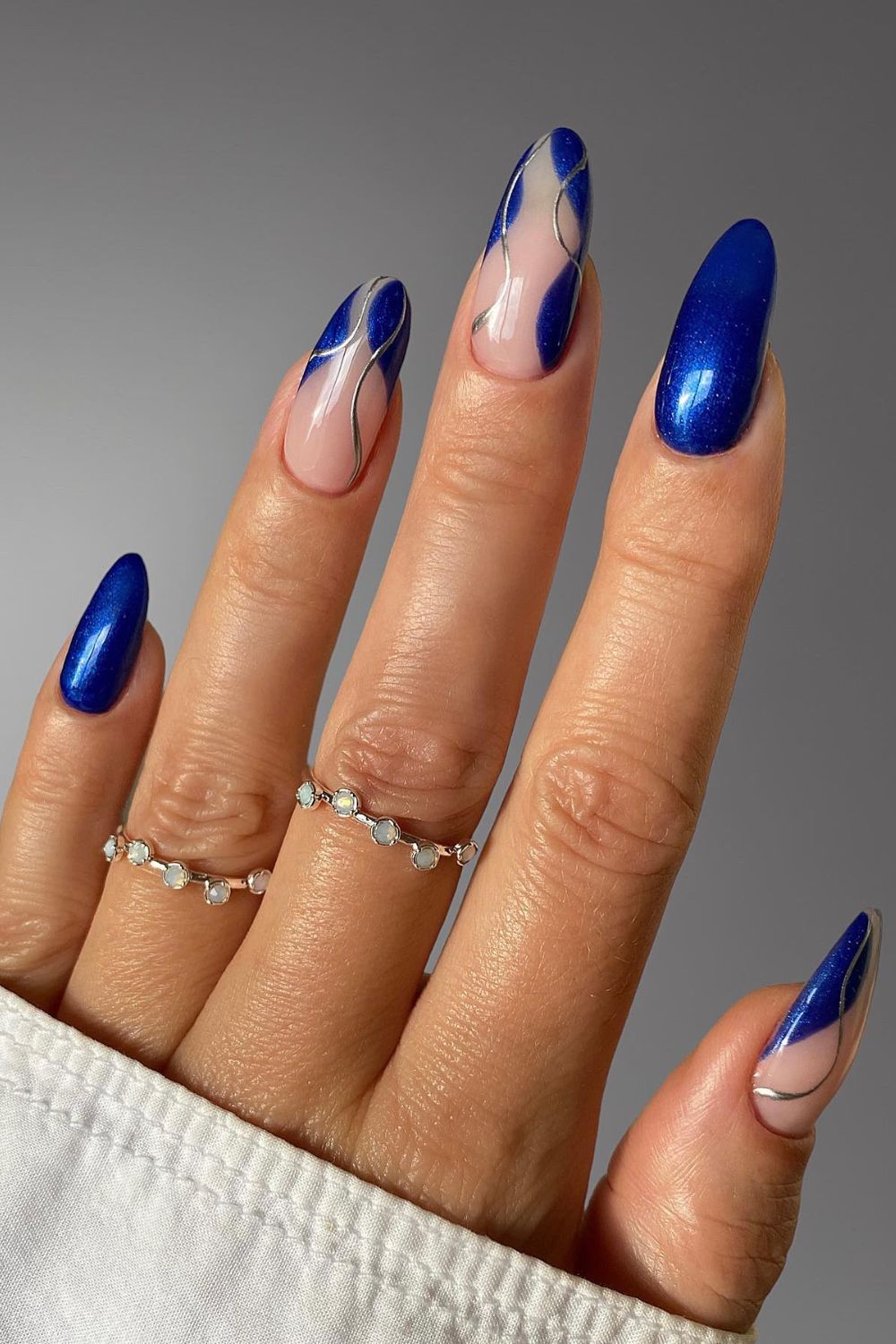 Long almond nails in deep cobalt-blue with a cat-eye shimmer, two nails with sheer silver swirl line art; real photography.