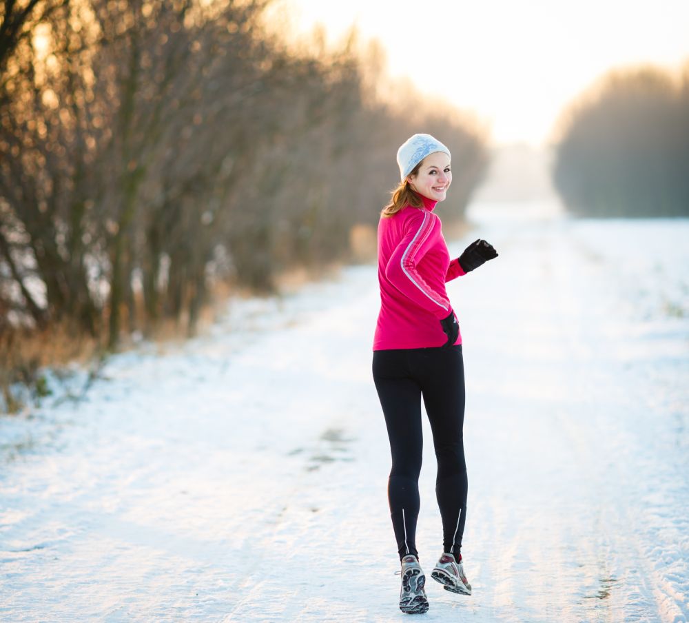Woman running on a snowy trail in pink jacket, black tights, beanie and gloves