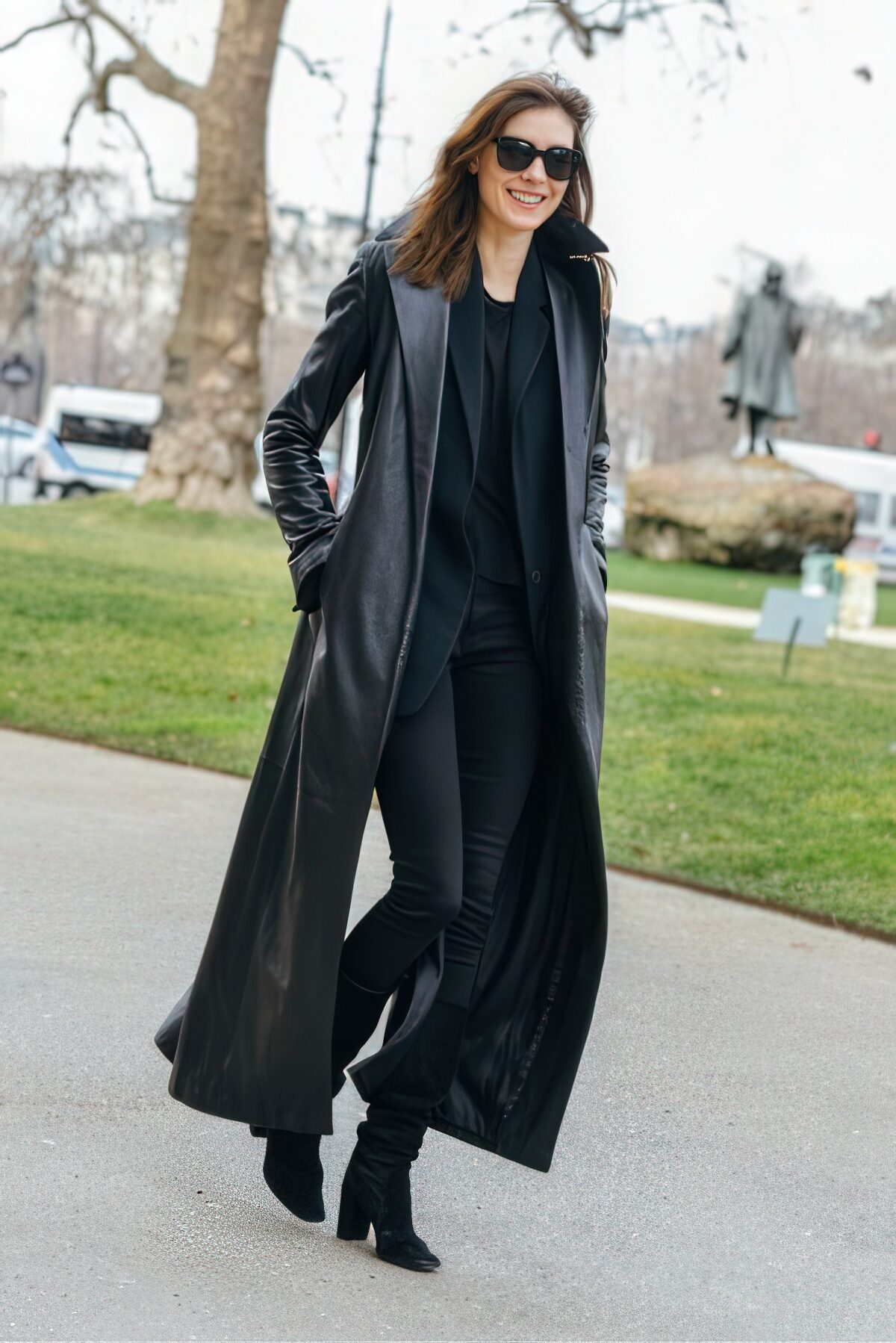 A black leather long coat over an all-black outfit of blazer, trousers and boots with sunglasses.