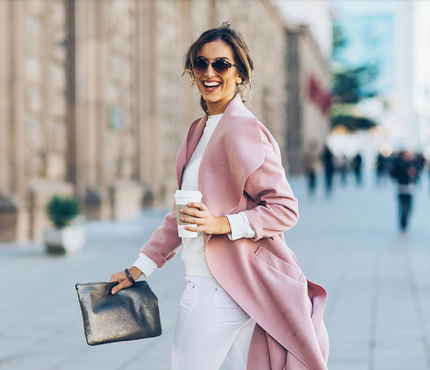 Woman wearing a dusty pink wrap coat with white jeans holding a metallic clutch