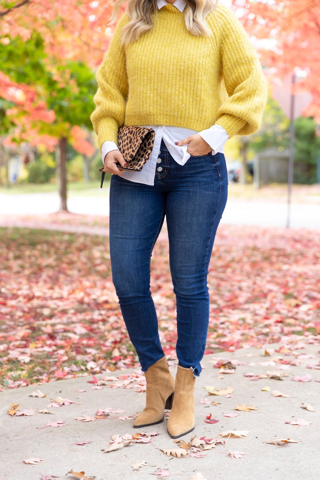 Woman wearing a mustard chunky knit sweater over a white collared shirt with dark jeans and tan ankle boots