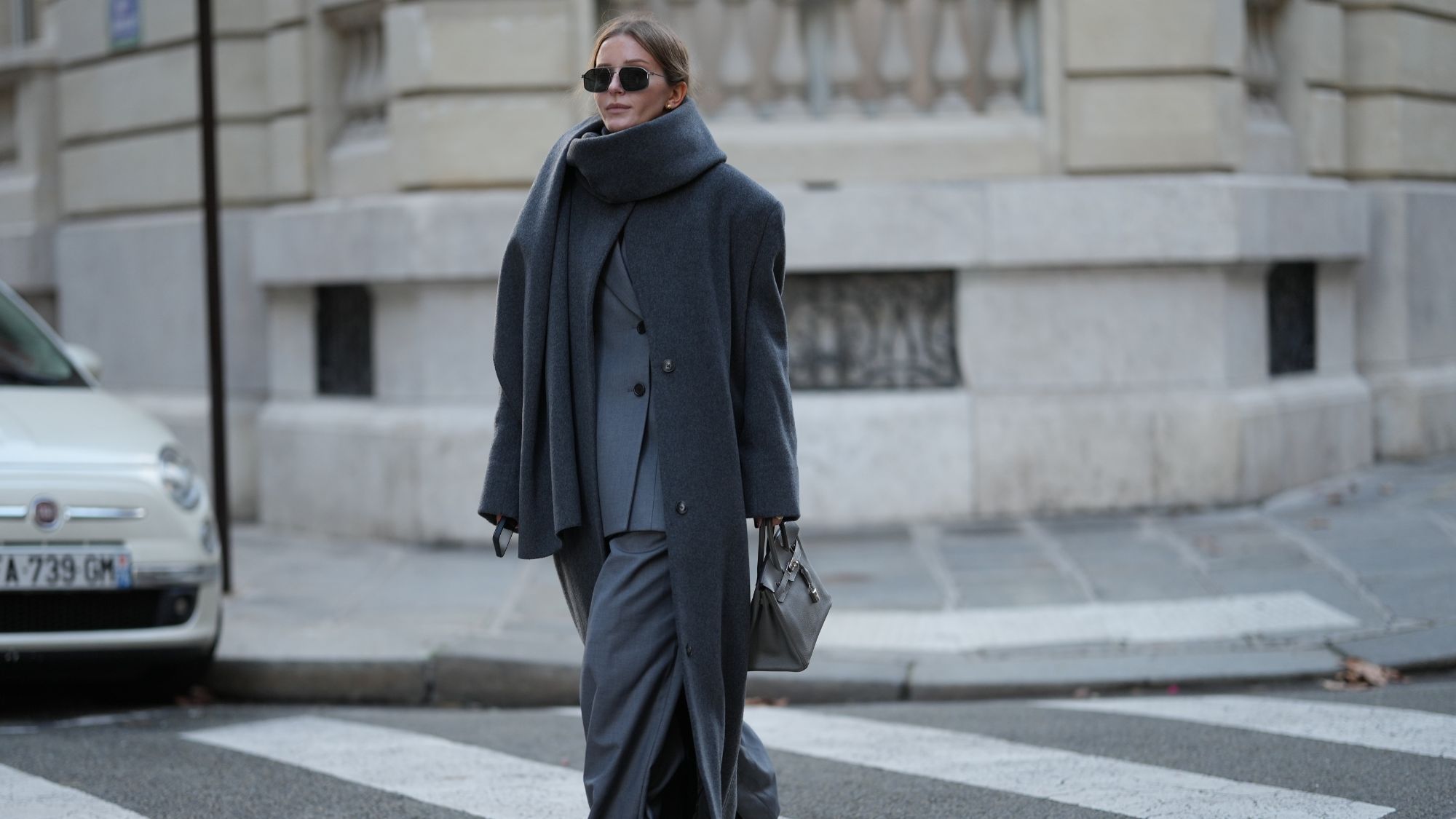 An oversized charcoal-grey wool coat with a draped scarf collar, worn over a grey blazer and wide-leg trousers with sunglasses and a structured bag.