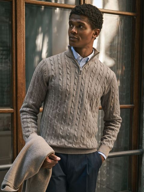 Man wearing a taupe cable-knit quarter-zip sweater over a striped shirt with dark trousers