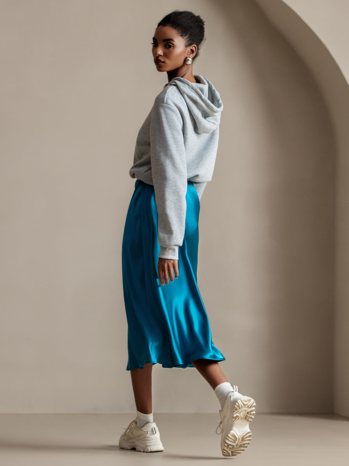 Woman in grey hoodie, jewel-blue satin midi skirt and chunky white sneakers