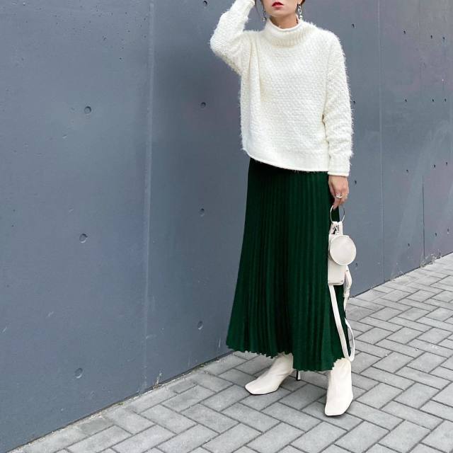 Woman wearing an emerald green pleated midi skirt, white textured knit sweater and white ankle boots