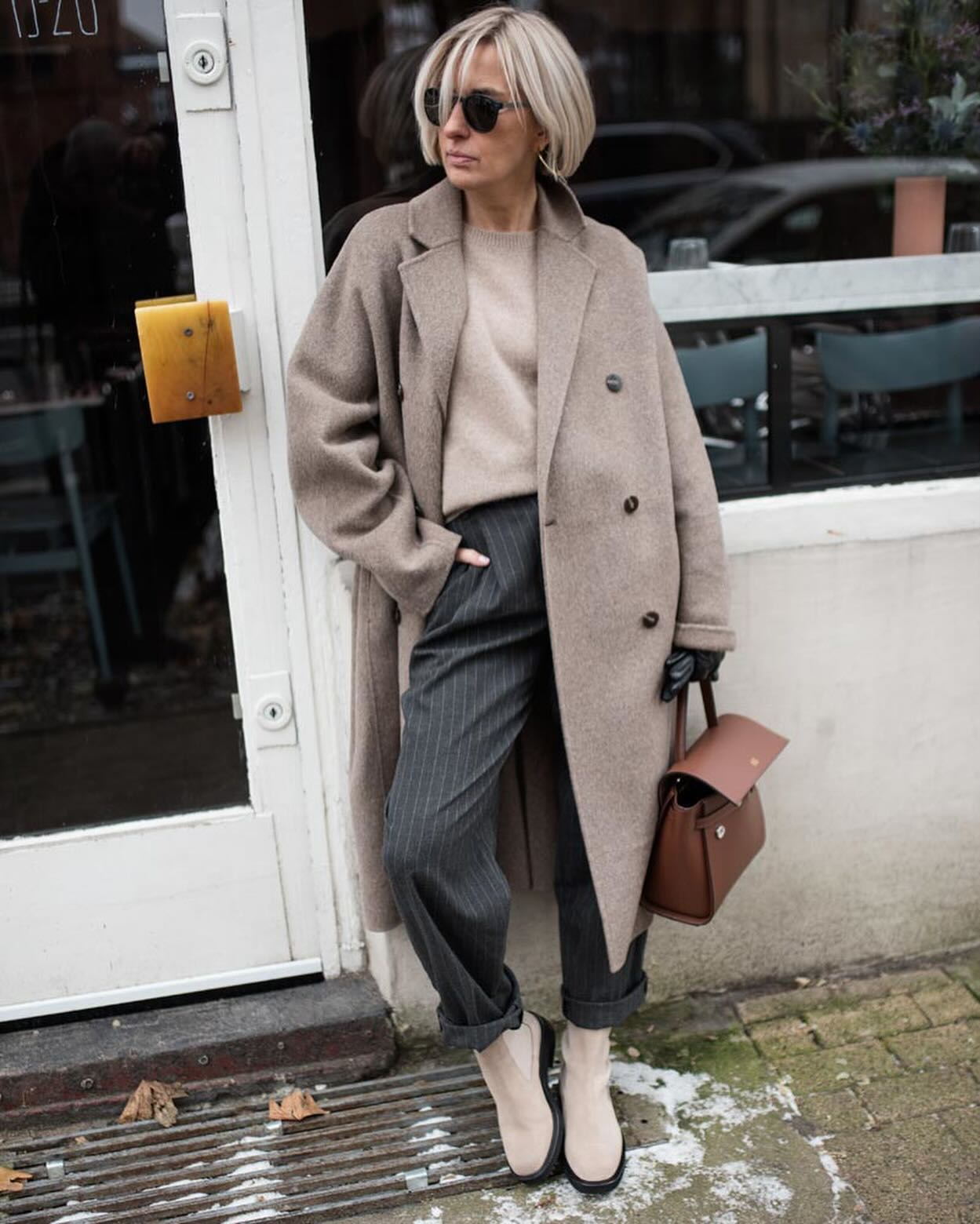 Woman in a camel wool coat, beige knit, pinstripe trousers and cream Chelsea boots on a snowy street
