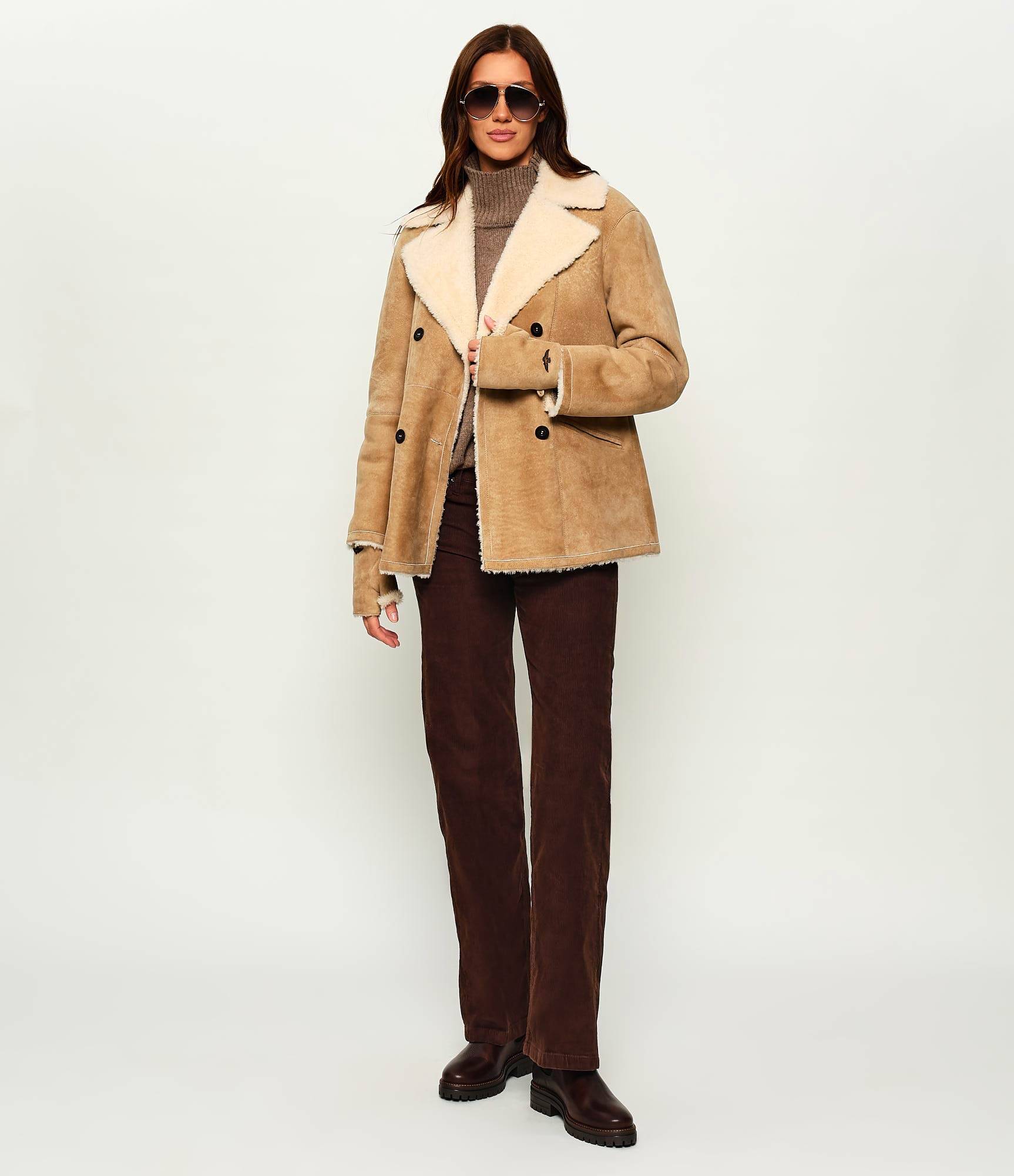 A camel double-breasted shearling coat with a cream shearling collar, worn over a taupe turtleneck and brown corduroy wide-leg trousers with chunky boots.