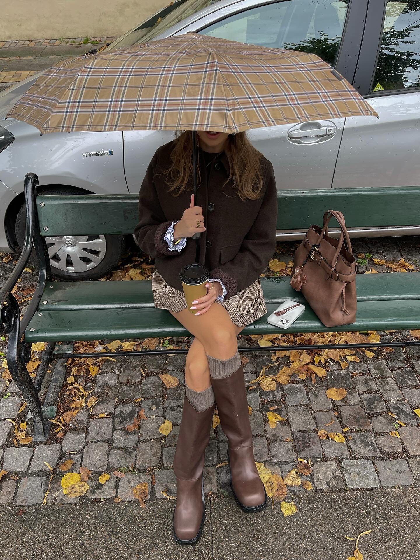 Woman in chocolate brown wool jacket, brown knee-high boots and tan leather tote, seated on a bench under a checked umbrella