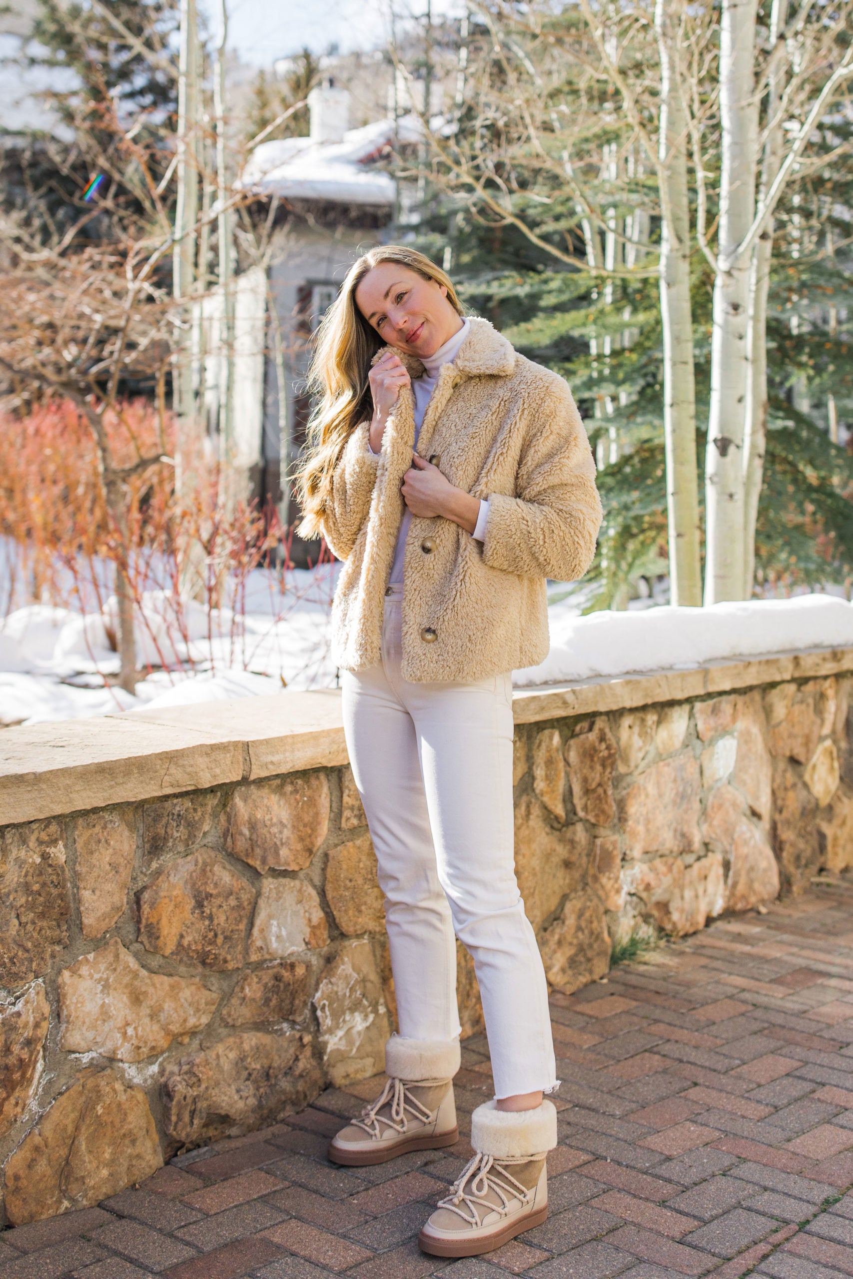 Woman in a beige teddy jacket, white jeans and fur-trim lace-up boots in a snowy alpine village