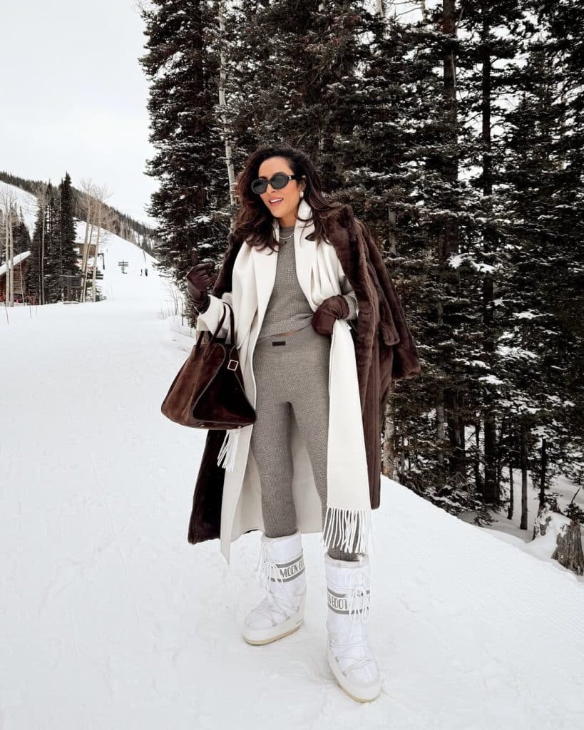 Woman in a brown faux-fur coat, grey knit set, white scarf and white moon boots on a snowy trail
