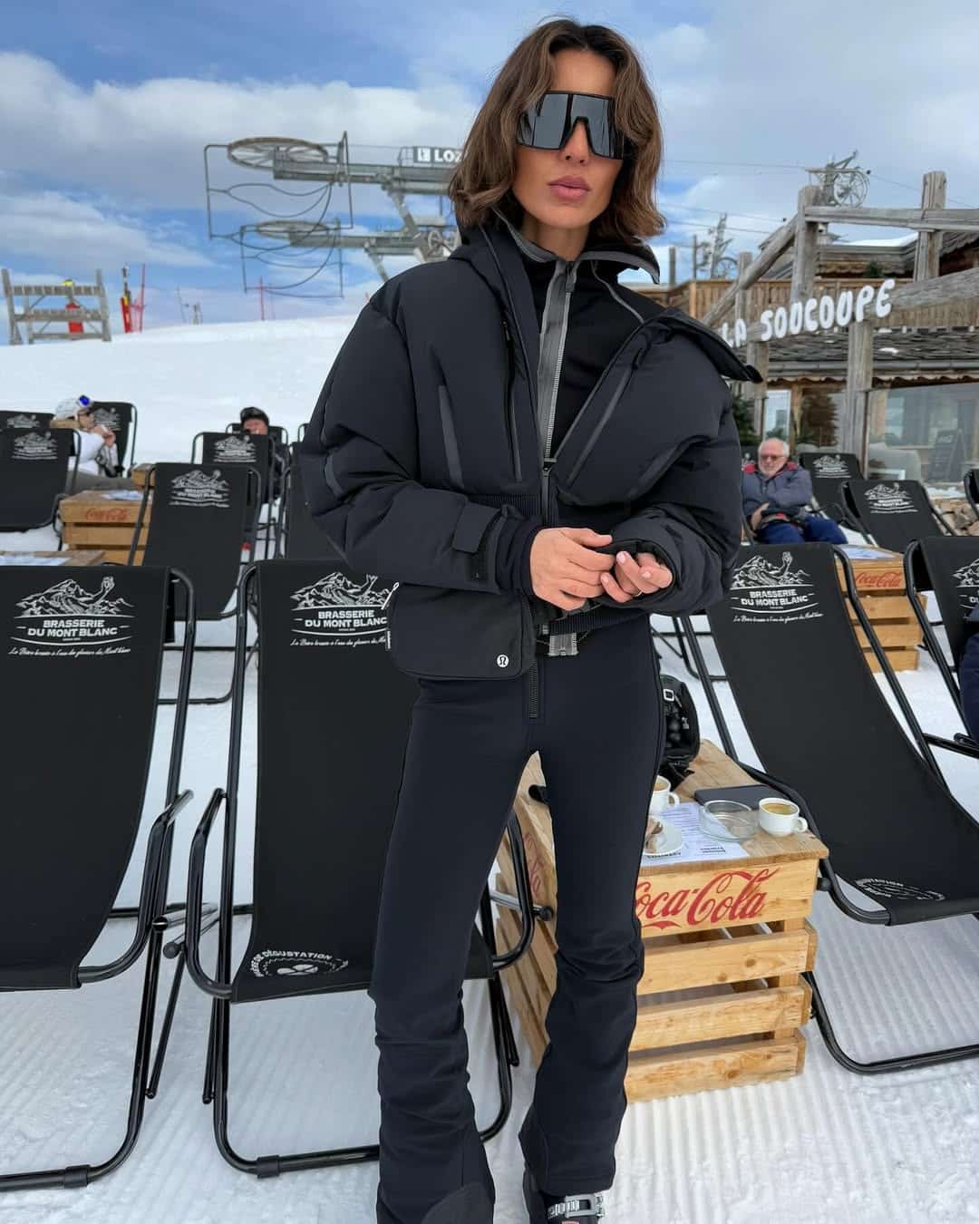 Woman in an all-black puffer ski set with large sunglasses standing at a snowy mountain restaurant terrace