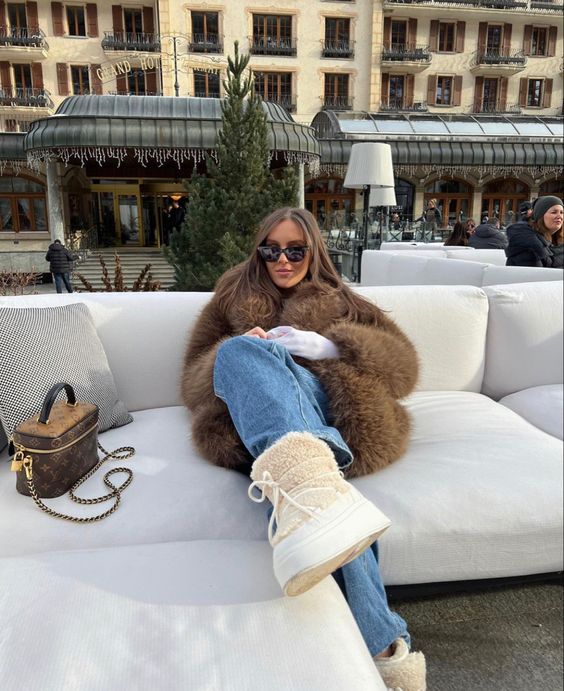 Woman in cream furry moon boots, chocolate brown faux-fur coat and blue jeans seated on an alpine terrace