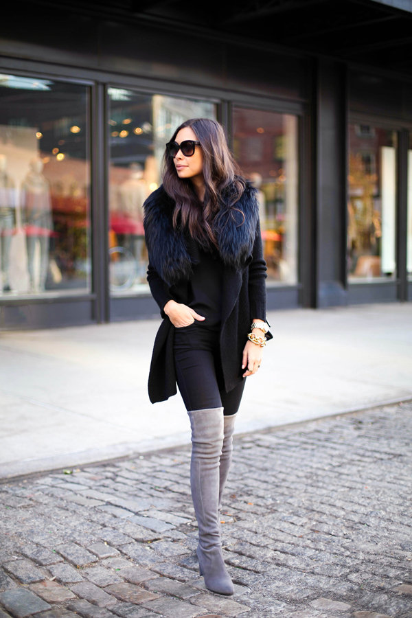 Woman in a black cardigan with a fur collar, black leggings and grey over-the-knee boots on a cobblestone street
