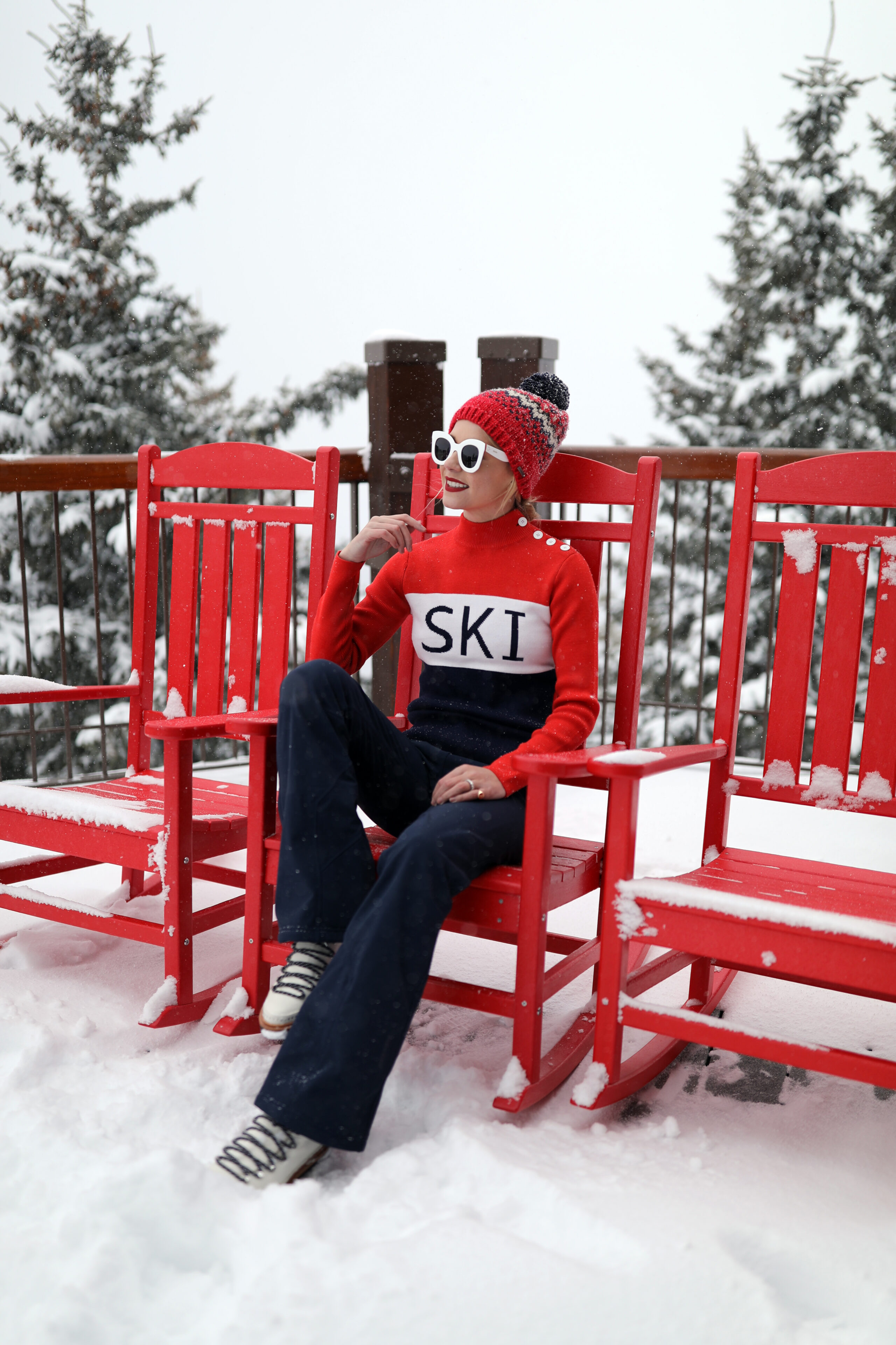 Woman in a red ski-lettered sweater, navy flare pants and red pom beanie on a snowy lodge deck