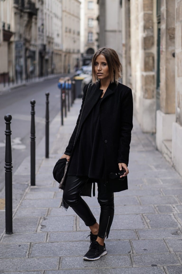 Fashion blogger wearing an all-black outfit: black peacoat, black top, black leather leggings and black sneakers on a Lyon street