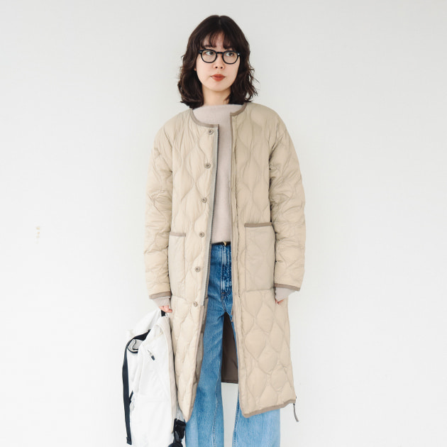 Long beige quilted coat worn over a cream knit and blue jeans with a white backpack
