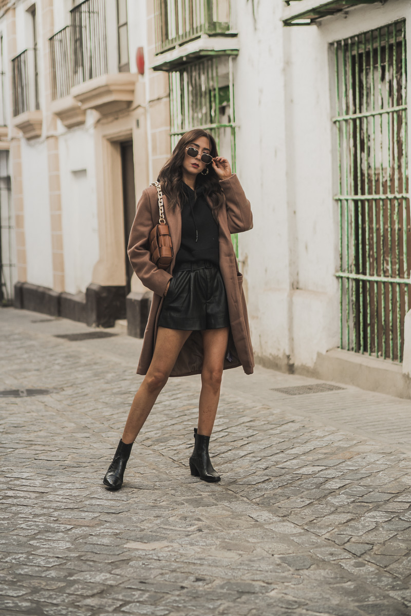 Woman in a camel coat over a black hoodie and black leather shorts with ankle boots