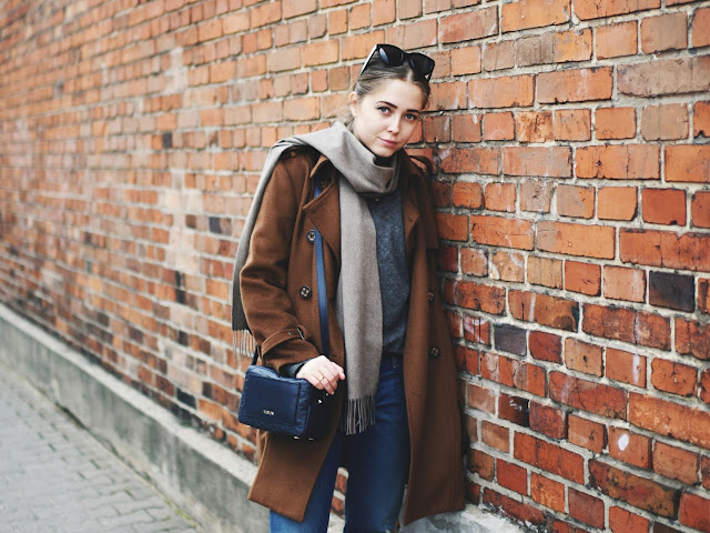 Woman in a cognac wool coat with a grey scarf, grey sweater, dark jeans and a navy bag