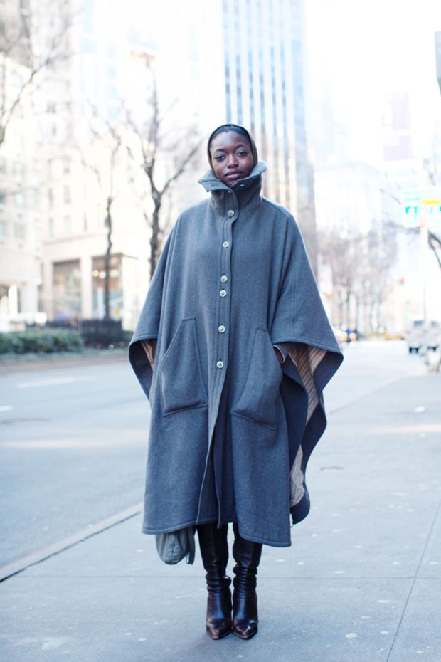 Woman in a gray button-front wool cape with a funnel neck and patch pockets, black tights, and black knee-high heeled boots on a city street