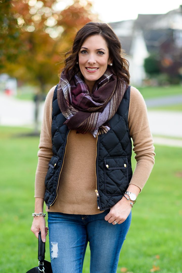 Woman wearing a black quilted vest over a camel sweater with a plaid scarf and jeans