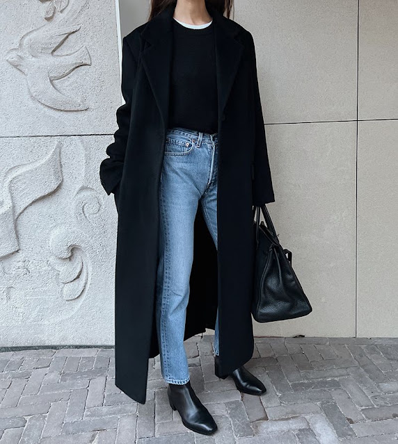 Woman wearing a black long coat with a black top, light-wash jeans, black ankle boots and a black bag