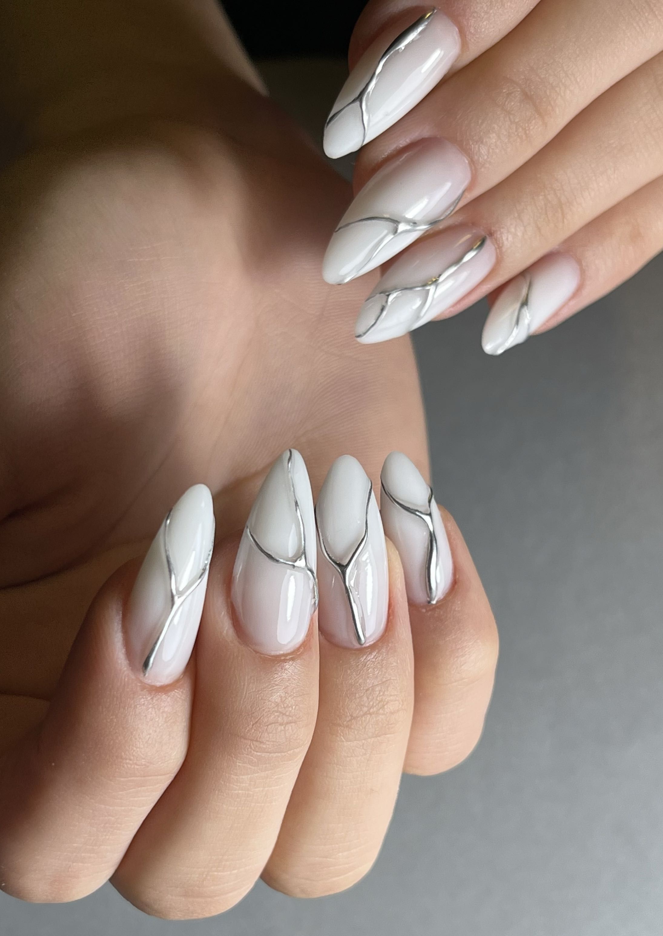 Milky winter-white almond nails decorated with thin silver chrome line art in flowing abstract patterns.