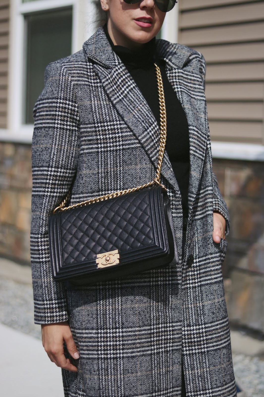 Woman in a gray plaid wool coat with a black turtleneck and a quilted chain-strap bag