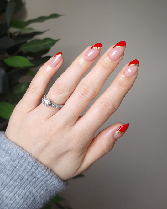 Medium almond nude nails with red French tips outlined in gold glitter