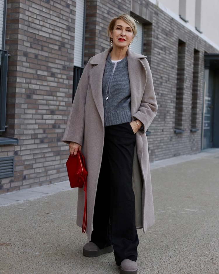 Woman in a long taupe wool coat with a grey knit, black wide-leg trousers and a red bag