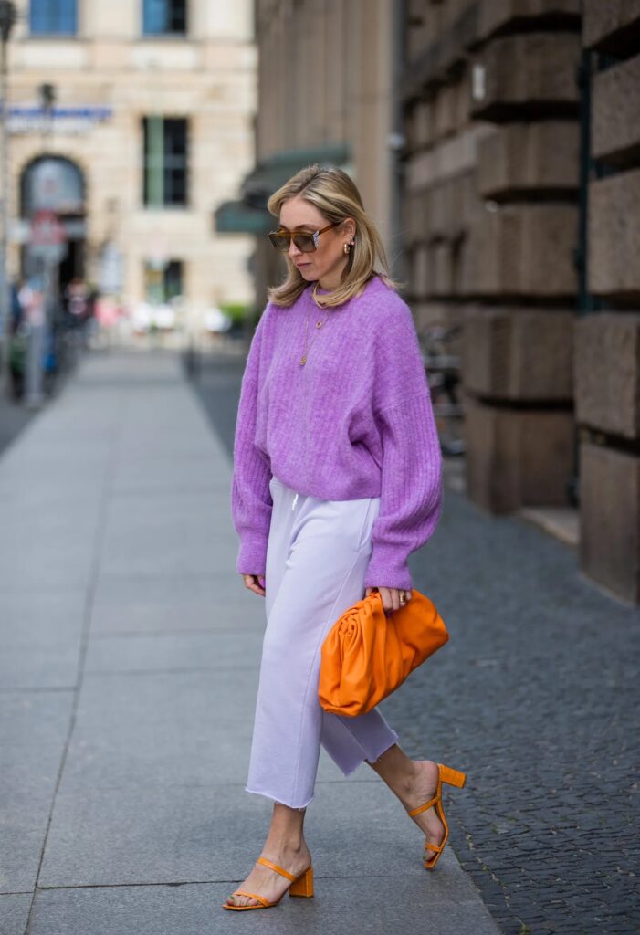 Woman in a lilac knit sweater and lilac wide-leg trousers with orange bag and sandals
