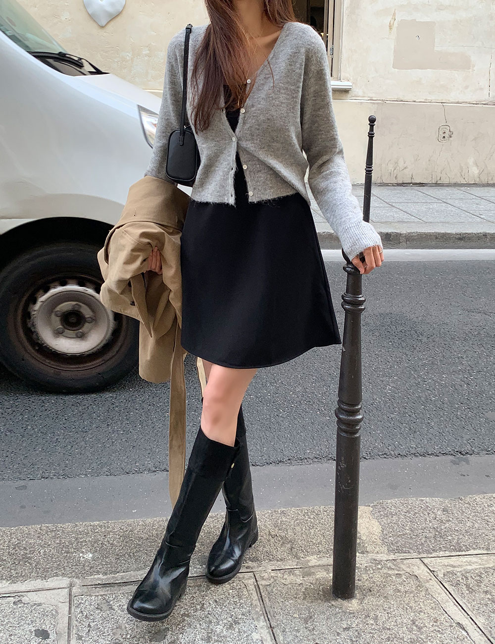 Woman wearing a grey V-neck cardigan over a black mini dress with black knee-high boots, holding a trench coat