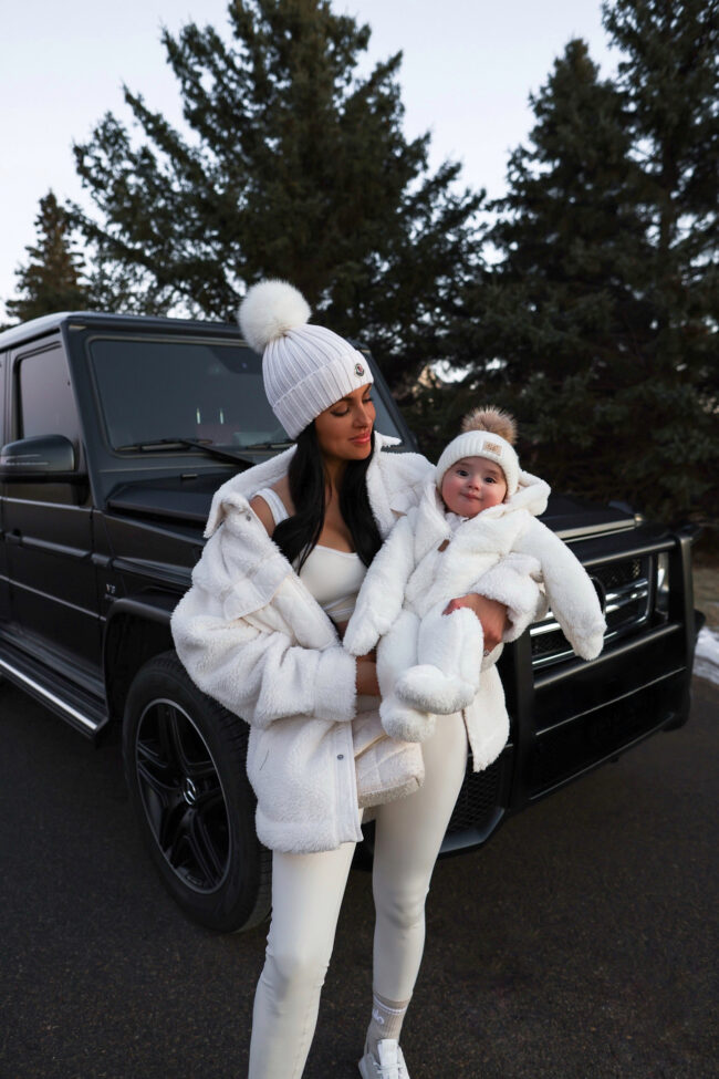 A mom and baby wearing matching white sherpa coats with knit pom hats in winter