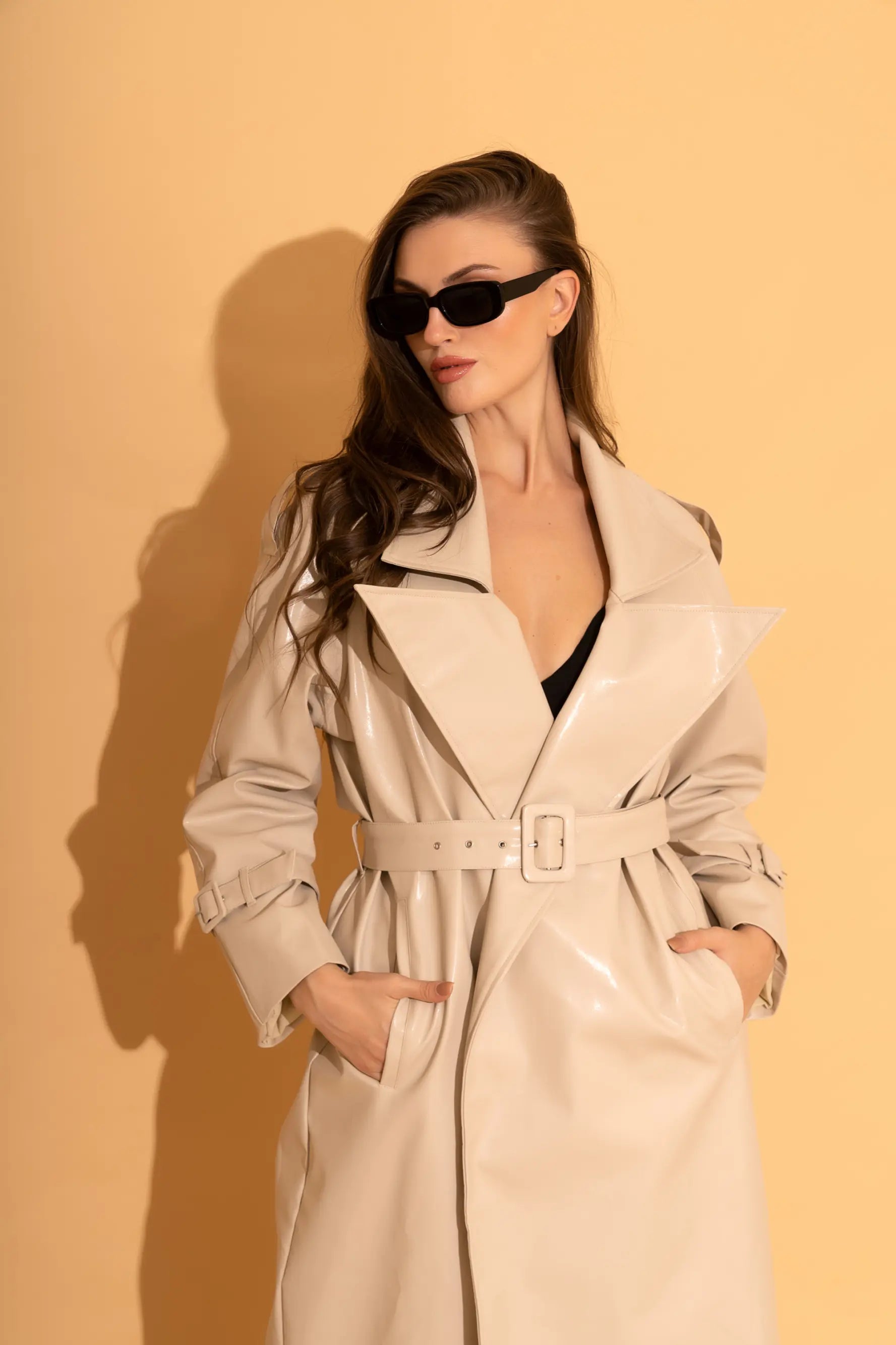 Woman in a cream patent faux-leather belted trench coat over a black top with dark sunglasses