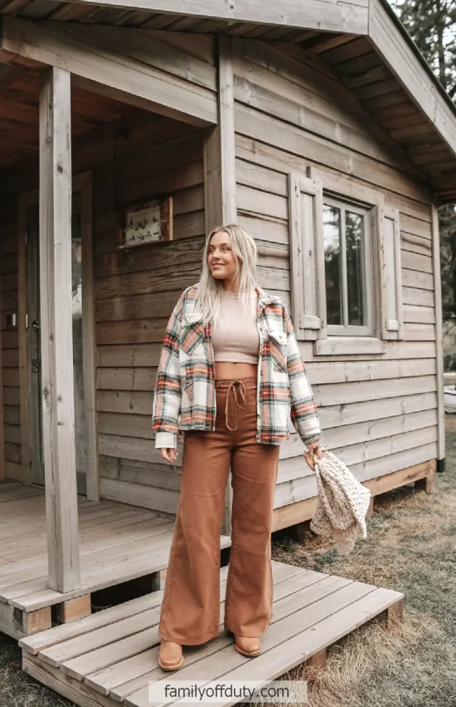 Woman in plaid shacket over a knit top with high-waisted wide-leg pants outside a wooden cabin
