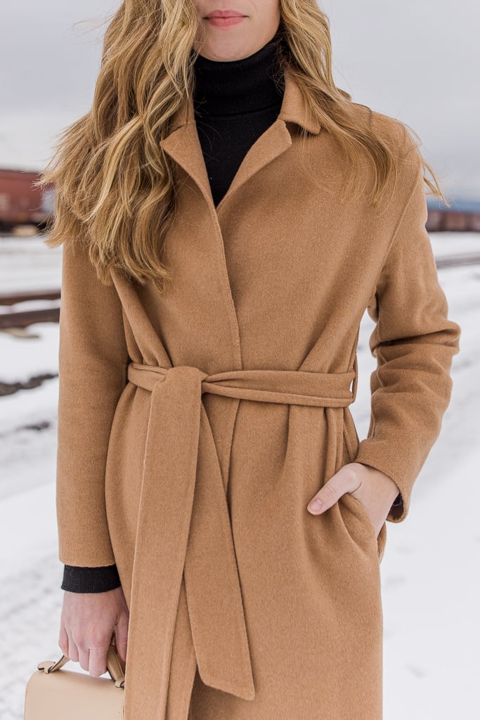 A camel belted wrap coat worn over a black turtleneck in a snowy setting.