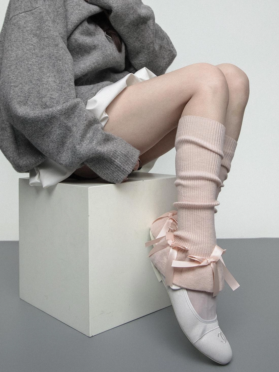 Pale pink leg warmers with satin ribbon bows worn with a grey oversized sweater and white ballet flats in a studio shot