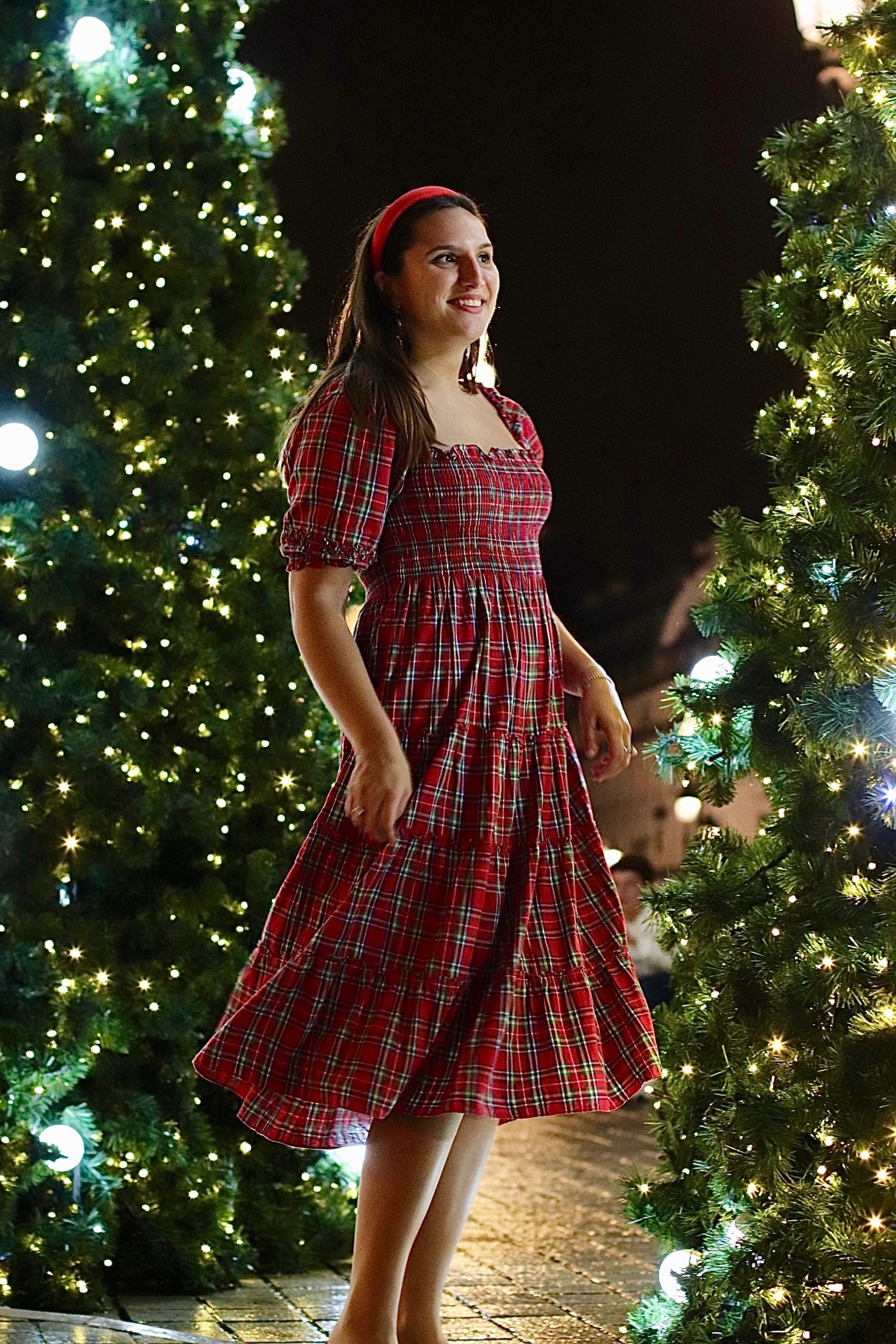 Woman in a red tartan smocked midi dress with puff sleeves and a red headband standing between illuminated Christmas trees at night