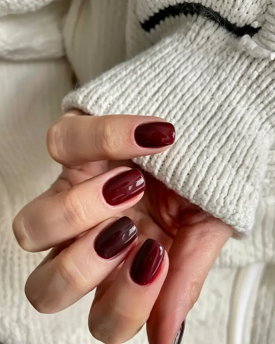 Short oval nails in deep cranberry wine polish against a white knit sweater
