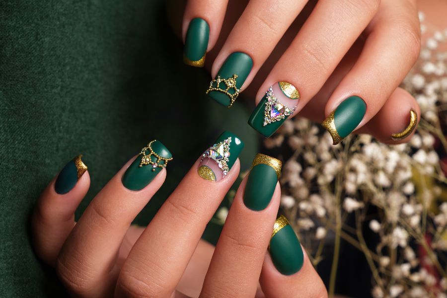 Medium almond nails in matte emerald green with gold glitter French tips and crystal embellishments