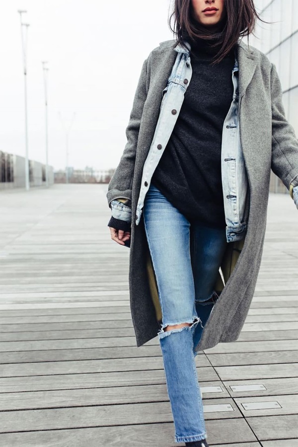 Woman layering a light-wash denim jacket under an oversized grey wool coat with a black sweater and ripped jeans