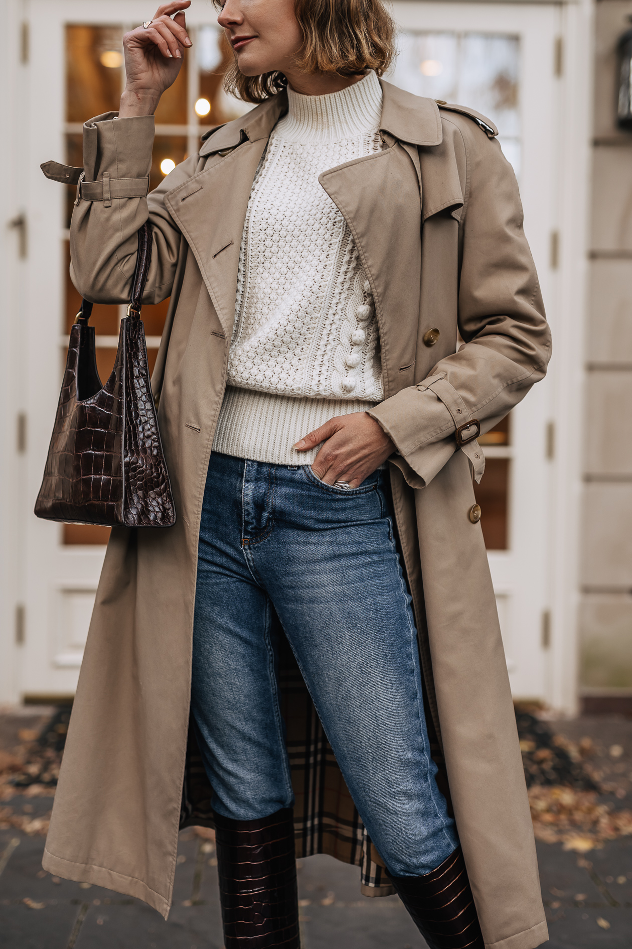 Woman wearing a beige trench coat over a cream chunky knit sweater with straight jeans and tall burgundy boots