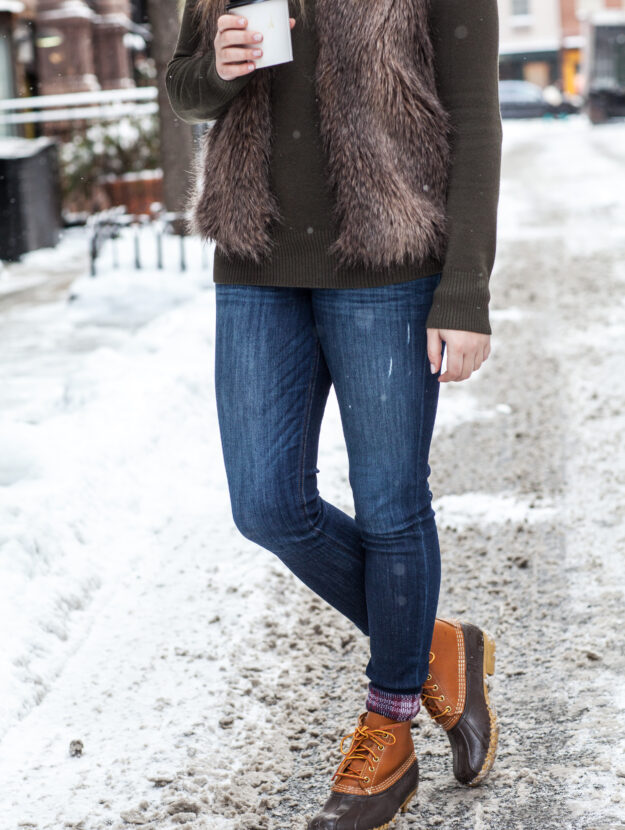 Woman in a faux-fur vest and jeans with tan duck boots holding a coffee in the snow