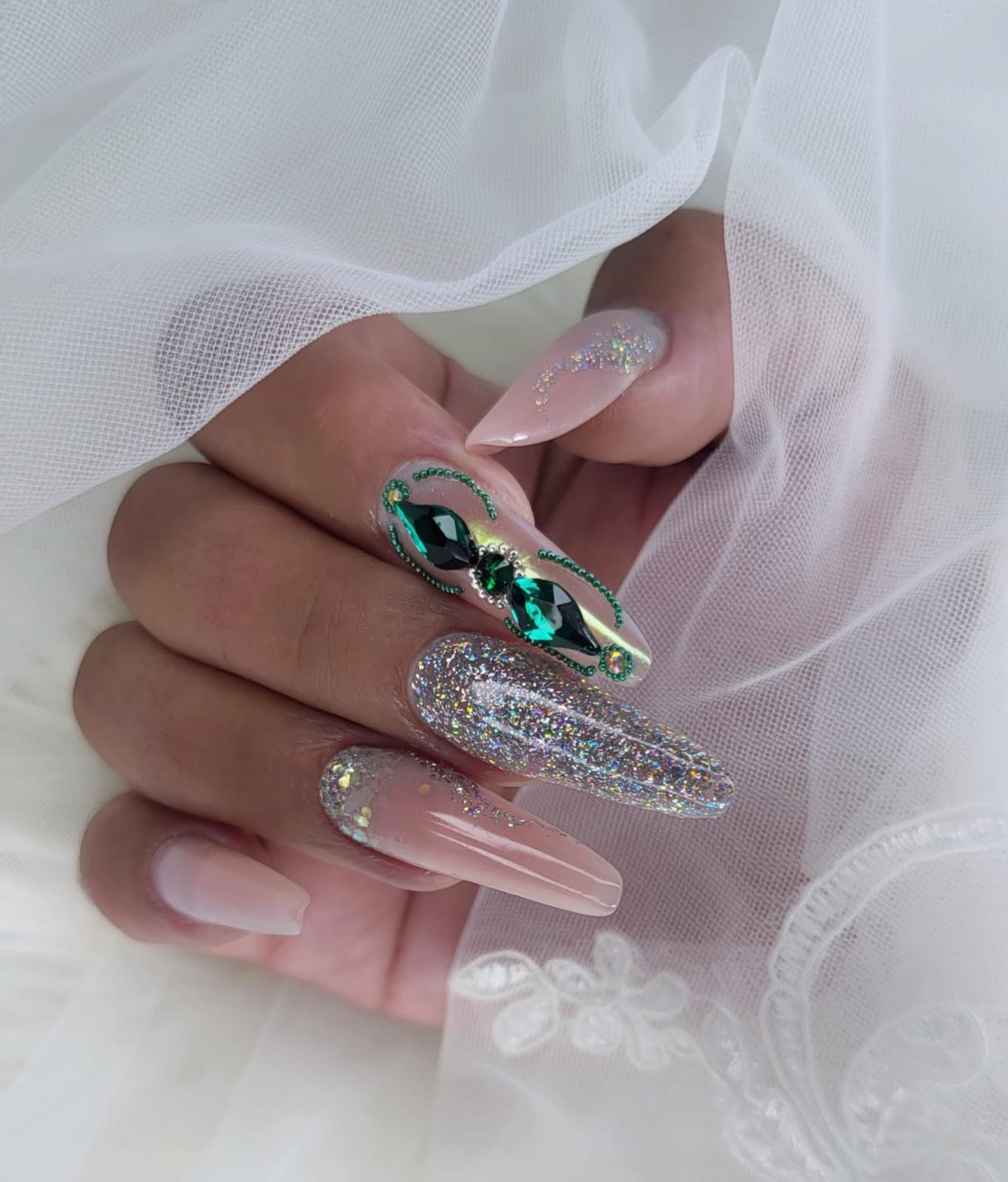 Long almond nails in sheer nude with large emerald-green crystal clusters and holographic glitter on accent nails